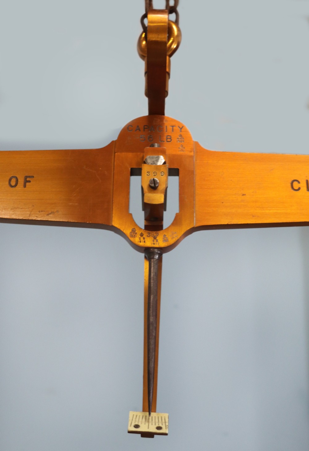 A large hanging brass beam balance scale featuring stamped lettering, suspended chain links, and two circular brass pans. View VI