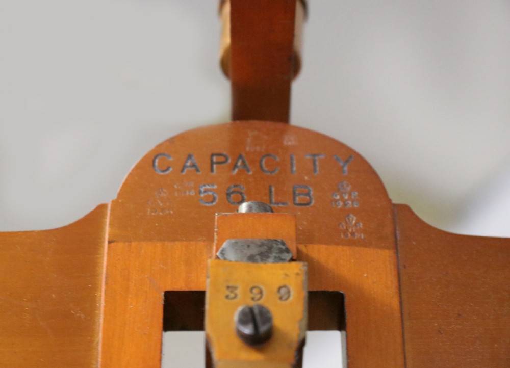 A large hanging brass beam balance scale featuring stamped lettering, suspended chain links, and two circular brass pans. View X