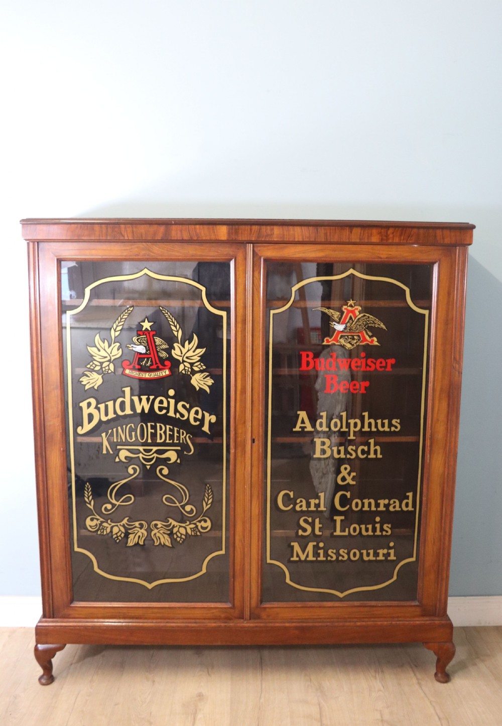 An early 20th-century walnut glazed display cabinet featuring two glass doors with gilded Budweiser advertising signwriting and cabriole legs.