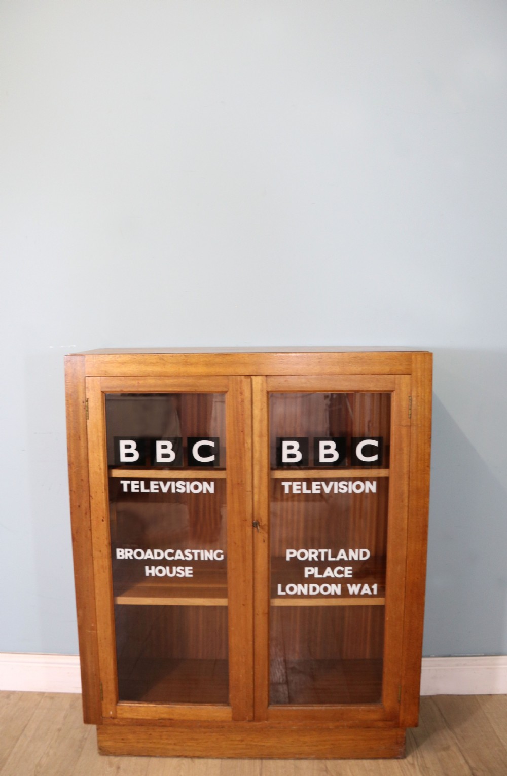 A 1930s light mahogany two-door glazed bookcase featuring white lettering on glass panels set over an enclosed wooden base.