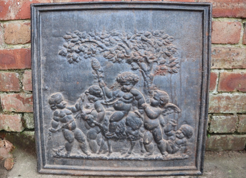 A square cast iron fireback featuring a raised relief scene of cherubs and putti beneath a leafy tree surrounded by a moulded border.