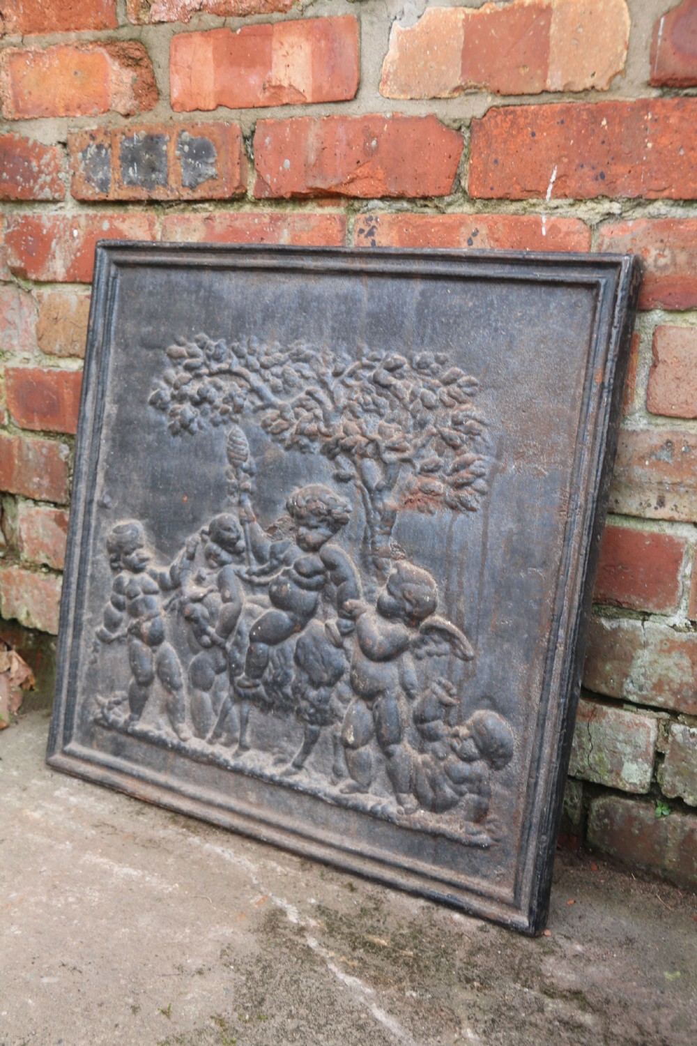 A square cast iron fireback featuring a raised relief scene of cherubs and putti beneath a leafy tree surrounded by a moulded border. View II