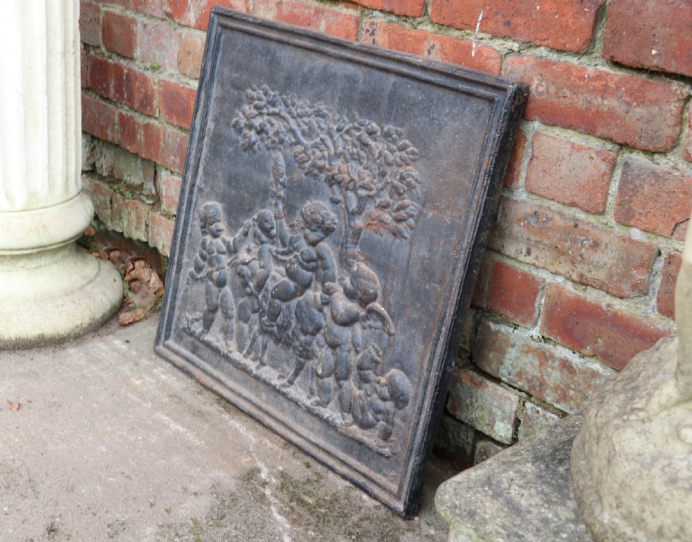 A square cast iron fireback featuring a raised relief scene of cherubs and putti beneath a leafy tree surrounded by a moulded border. View III