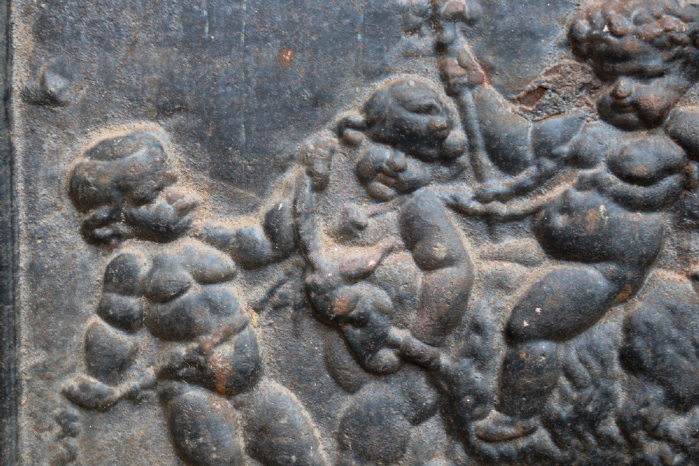 A square cast iron fireback featuring a raised relief scene of cherubs and putti beneath a leafy tree surrounded by a moulded border. View IV