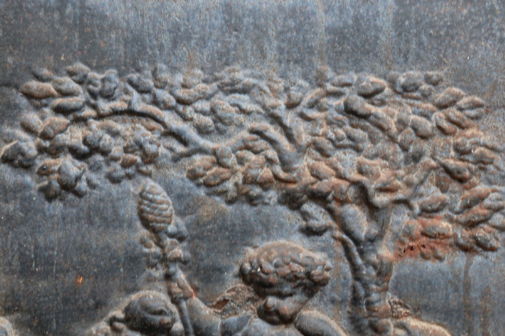 A square cast iron fireback featuring a raised relief scene of cherubs and putti beneath a leafy tree surrounded by a moulded border. View V