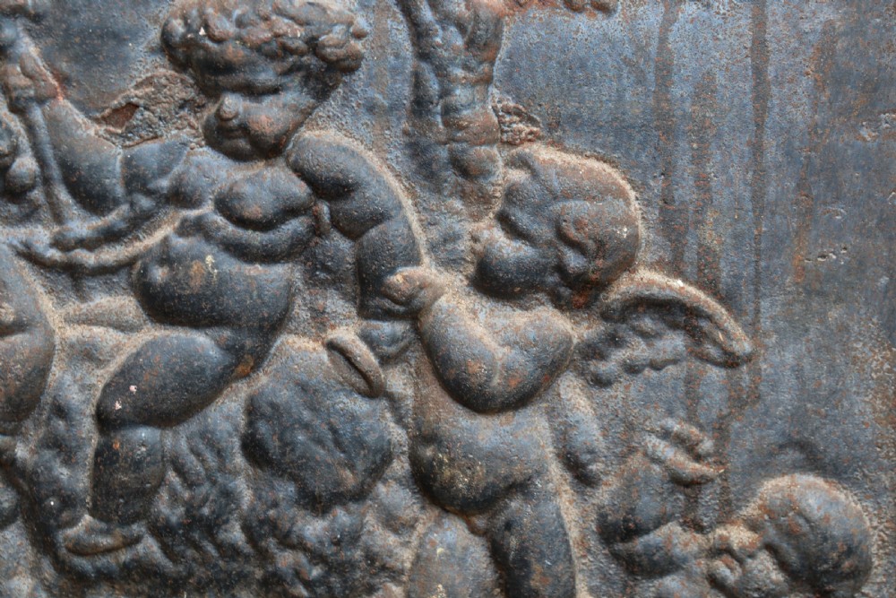 A square cast iron fireback featuring a raised relief scene of cherubs and putti beneath a leafy tree surrounded by a moulded border. View VI
