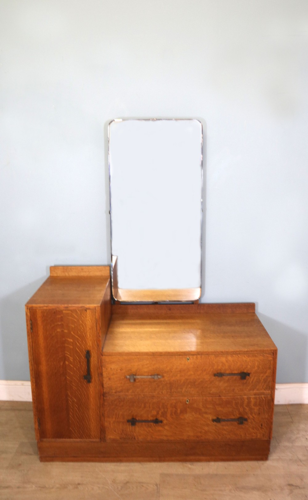 An asymmetrical golden oak dressing chest with a left-hand side cupboard, two drawers on the right, metal handles, and an off-centre tall mirror.