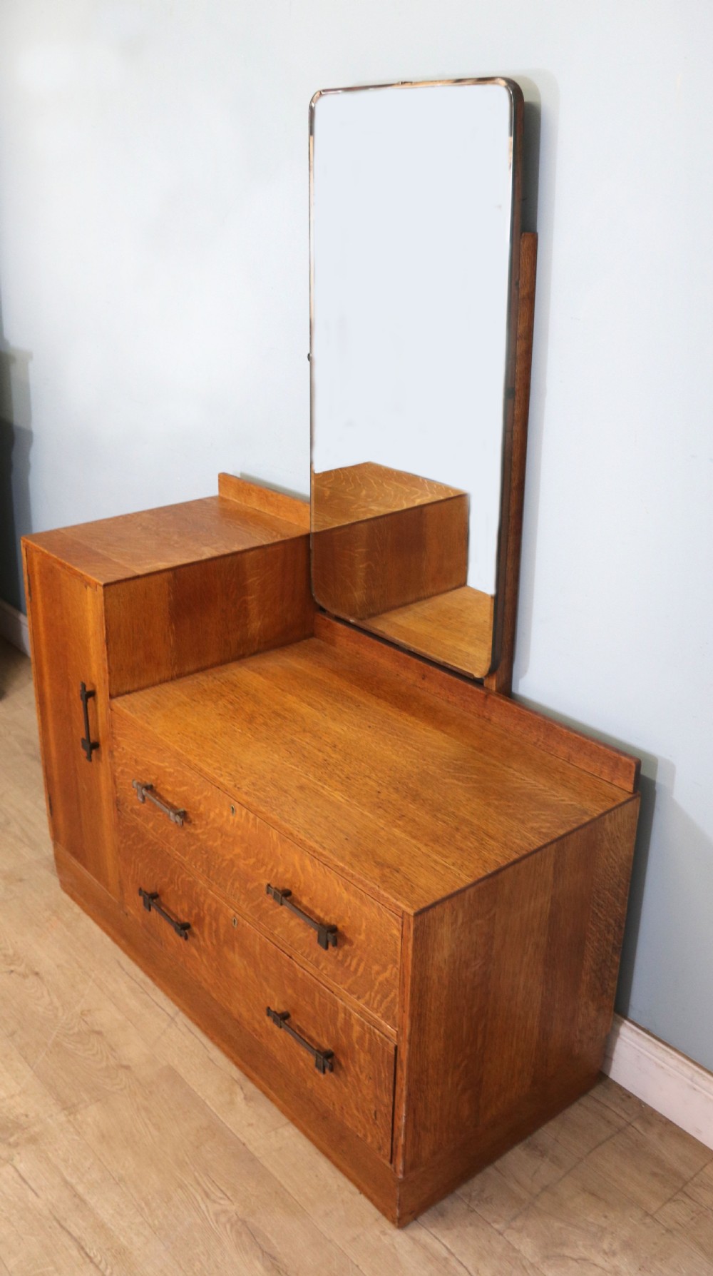 An asymmetrical golden oak dressing chest with a left-hand side cupboard, two drawers on the right, metal handles, and an off-centre tall mirror. View III