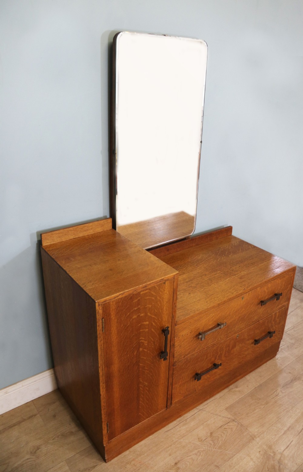 An asymmetrical golden oak dressing chest with a left-hand side cupboard, two drawers on the right, metal handles, and an off-centre tall mirror. View IV