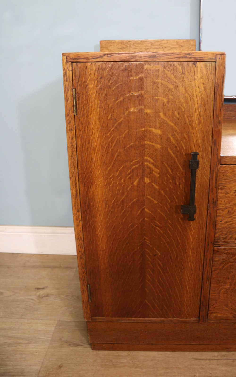 An asymmetrical golden oak dressing chest with a left-hand side cupboard, two drawers on the right, metal handles, and an off-centre tall mirror. View XIX