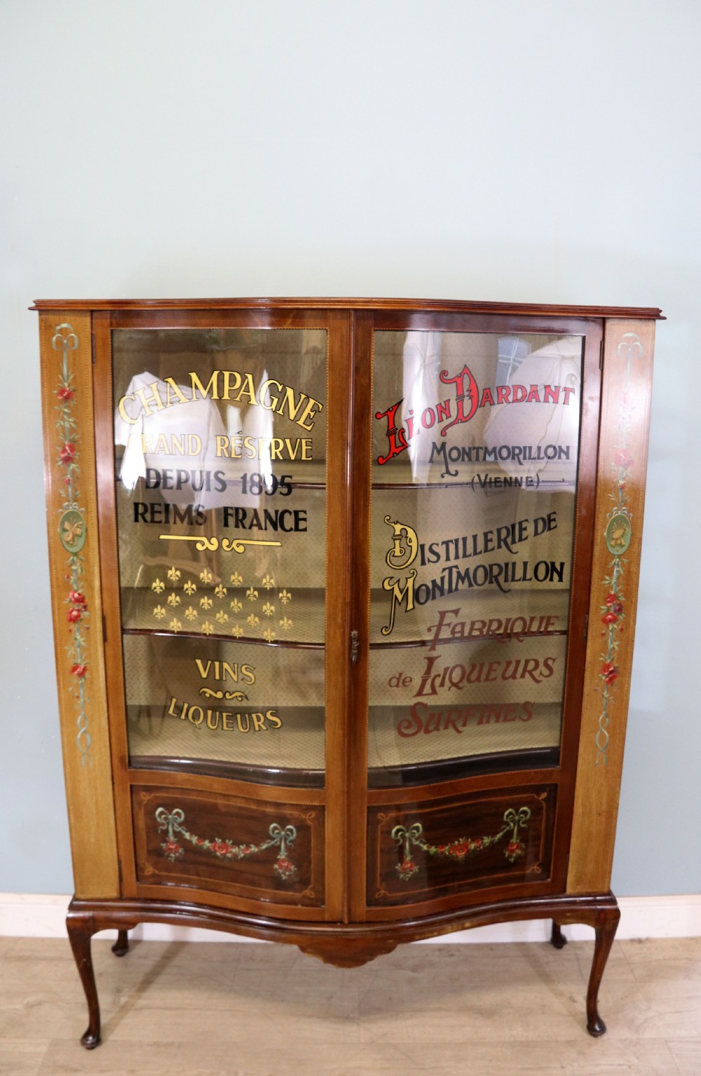 An Edwardian mahogany and satinwood display cabinet featuring curved glass doors decorated with French text and painted floral motifs, resting on slender cabriole legs.
