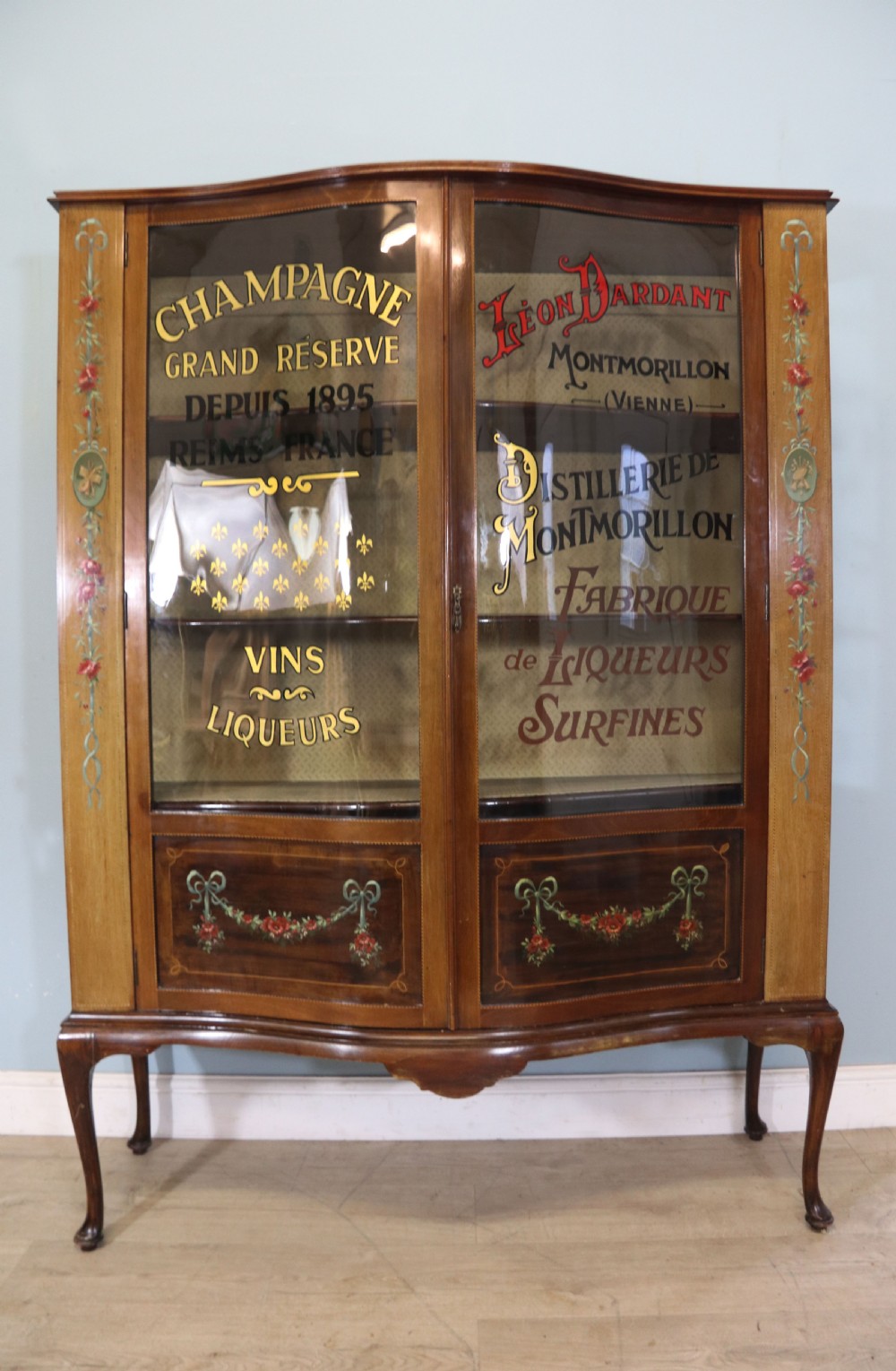 An Edwardian mahogany and satinwood display cabinet featuring curved glass doors decorated with French text and painted floral motifs, resting on slender cabriole legs. View II
