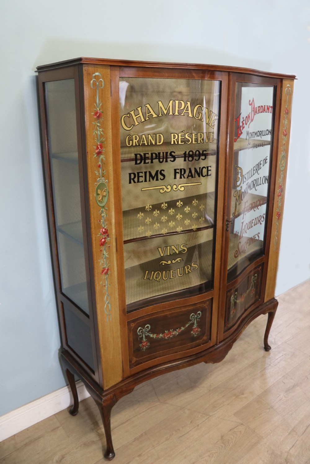 An Edwardian mahogany and satinwood display cabinet featuring curved glass doors decorated with French text and painted floral motifs, resting on slender cabriole legs. View IV