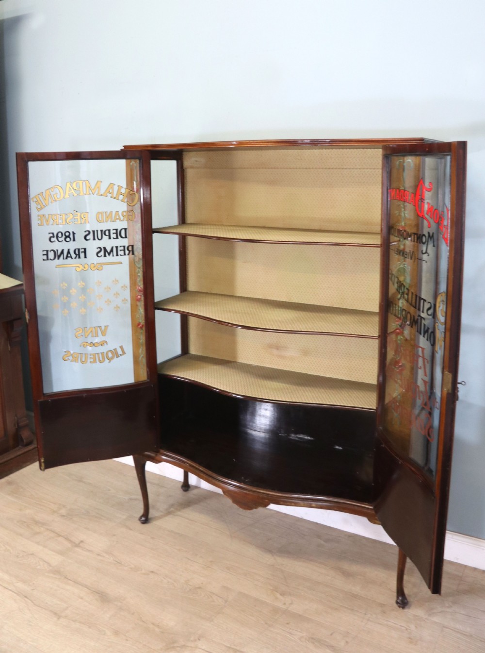 An Edwardian mahogany and satinwood display cabinet featuring curved glass doors decorated with French text and painted floral motifs, resting on slender cabriole legs. View VI