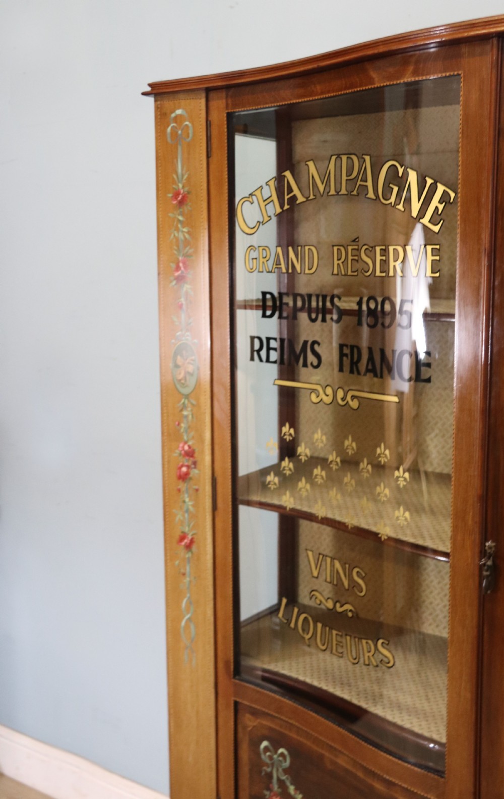 An Edwardian mahogany and satinwood display cabinet featuring curved glass doors decorated with French text and painted floral motifs, resting on slender cabriole legs. View X