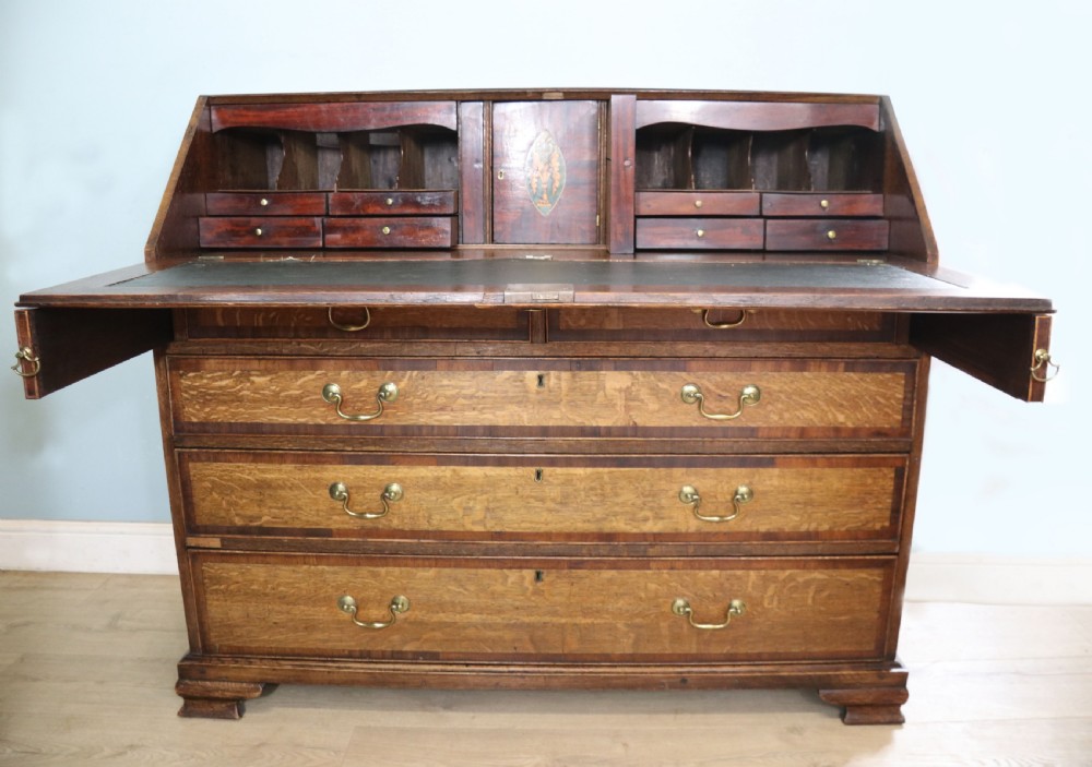 An open George III oak bureau featuring mahogany crossbanding, brass swan-neck handles, pull-out lopers, and a fitted interior with pigeonholes.