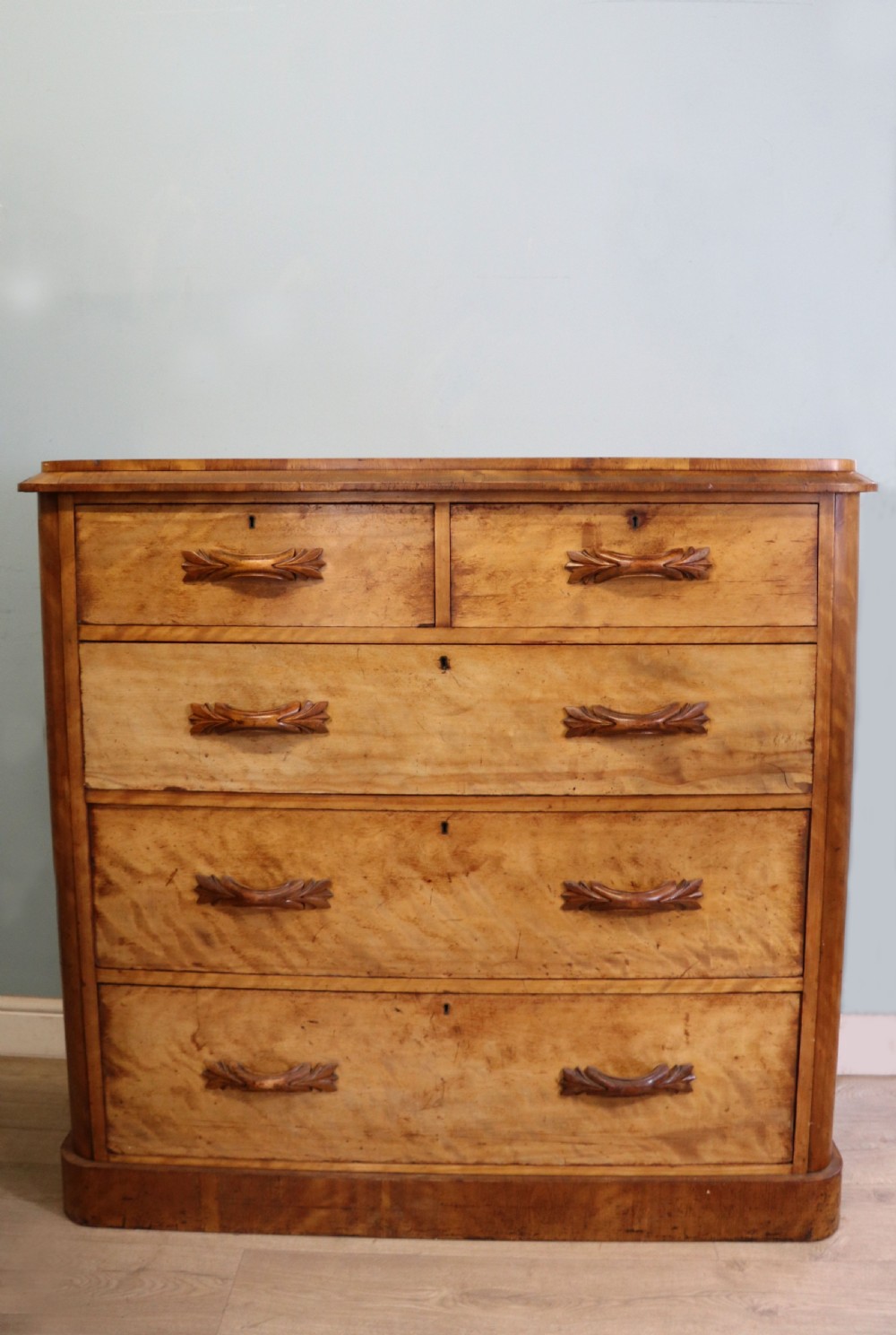 A Victorian light wood chest of drawers with two short drawers over three long drawers, featuring carved leaf handles and a solid plinth base.
