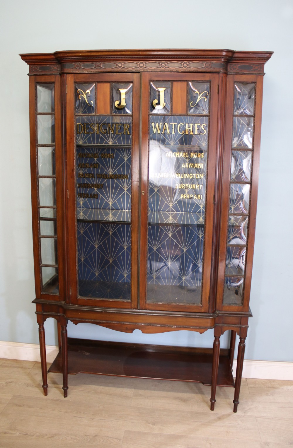 A tall mahogany glazed display cabinet featuring two central glass doors, glazed sides, turned legs, a lower undertier shelf, and gilded gold lettering inside.