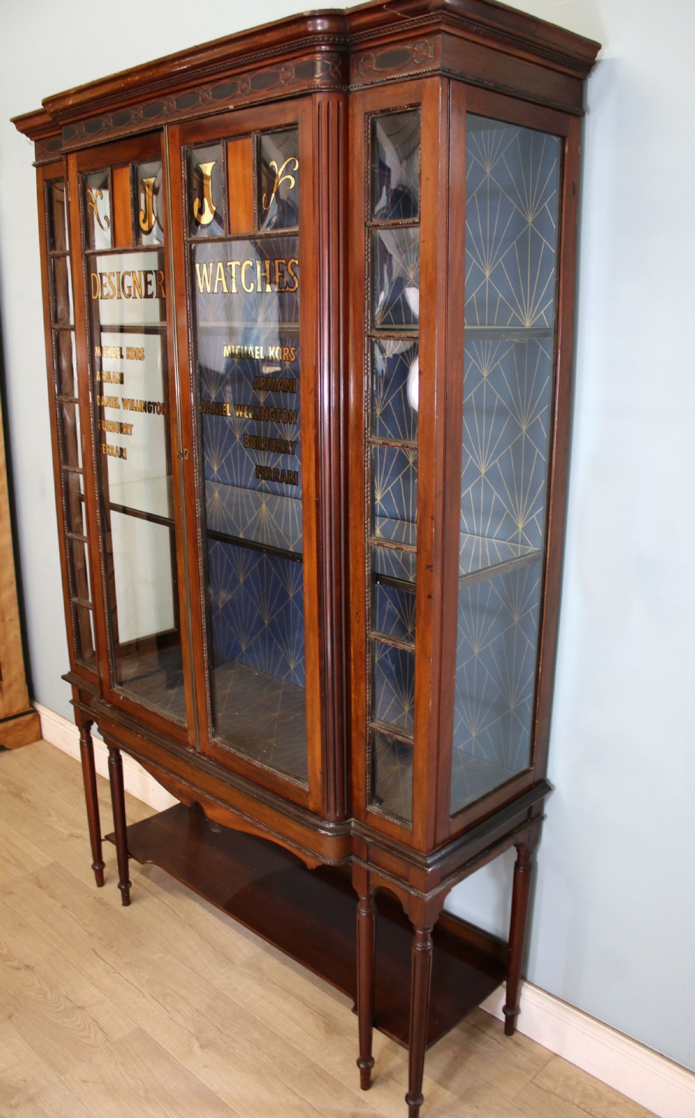 A tall mahogany glazed display cabinet featuring two central glass doors, glazed sides, turned legs, a lower undertier shelf, and gilded gold lettering inside. View III