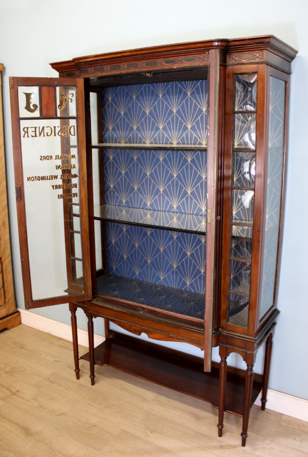A tall mahogany glazed display cabinet featuring two central glass doors, glazed sides, turned legs, a lower undertier shelf, and gilded gold lettering inside. View IV