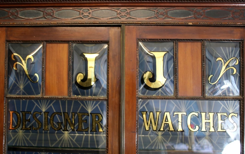 A tall mahogany glazed display cabinet featuring two central glass doors, glazed sides, turned legs, a lower undertier shelf, and gilded gold lettering inside. View VI