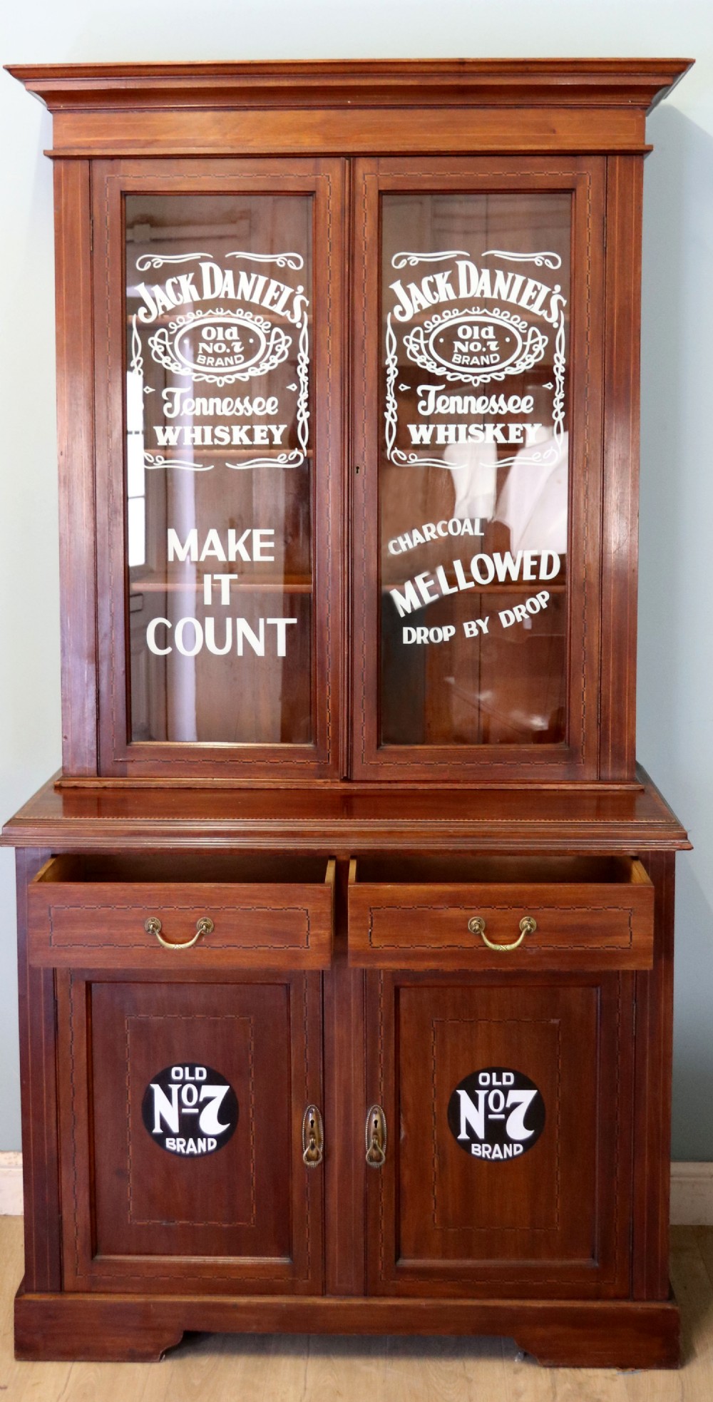 A late Victorian warm brown wooden cabinet featuring a glazed upper display section with white Jack Daniels typography and two lower panelled doors. View II