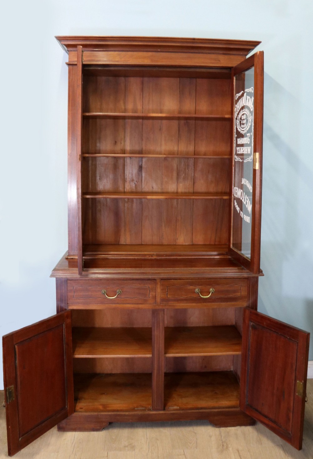 A late Victorian warm brown wooden cabinet featuring a glazed upper display section with white Jack Daniels typography and two lower panelled doors. View III