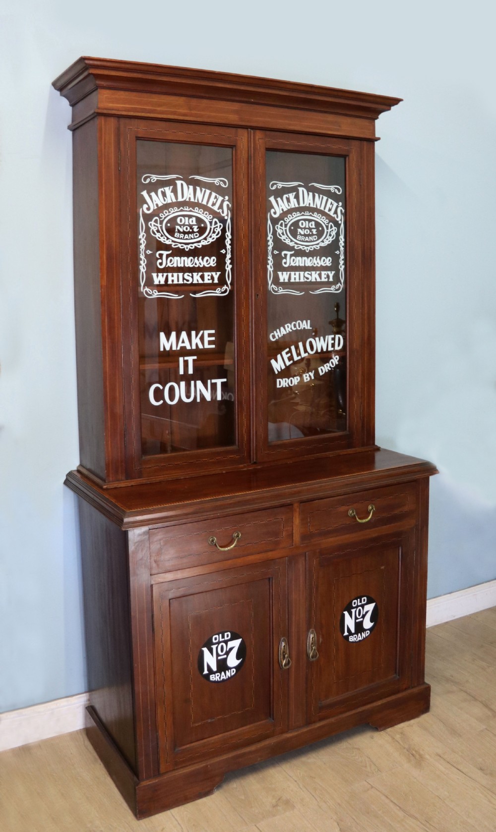 A late Victorian warm brown wooden cabinet featuring a glazed upper display section with white Jack Daniels typography and two lower panelled doors. View IV