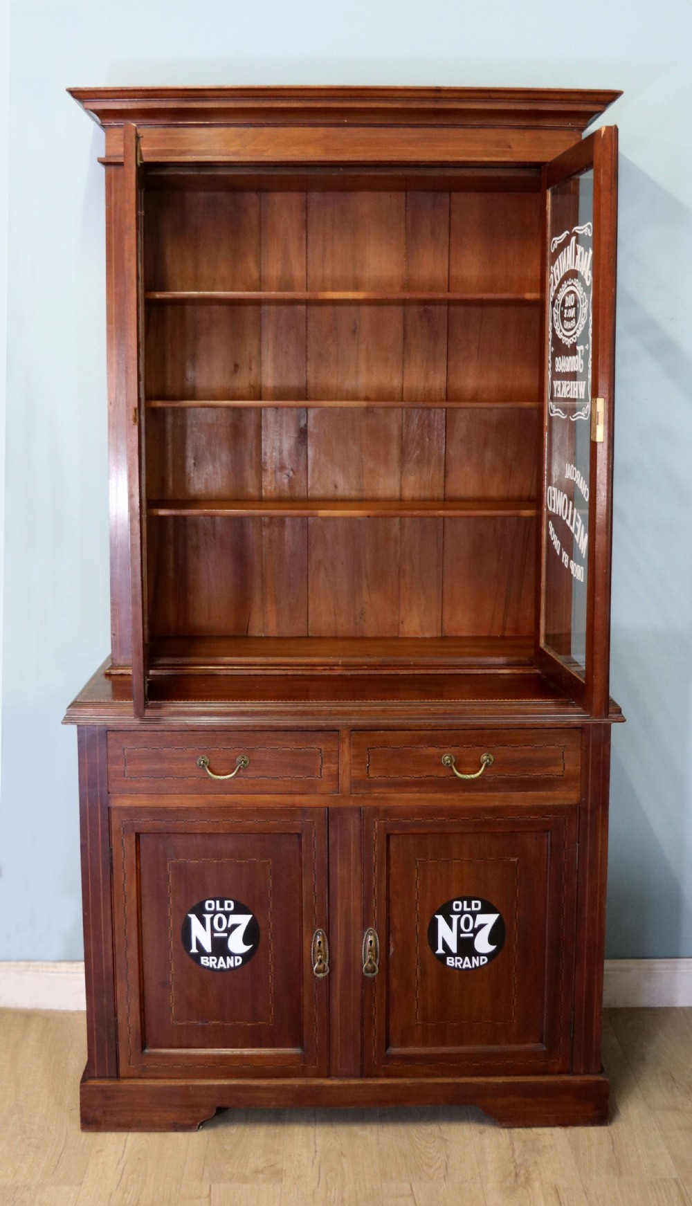 A late Victorian warm brown wooden cabinet featuring a glazed upper display section with white Jack Daniels typography and two lower panelled doors. View V