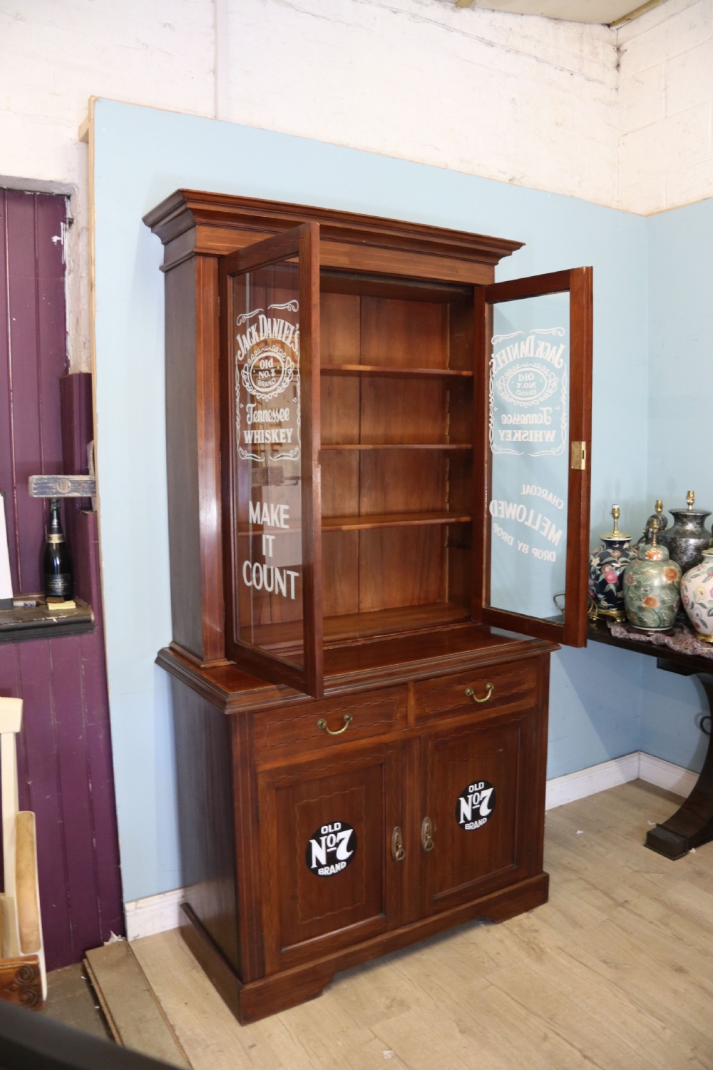 A late Victorian warm brown wooden cabinet featuring a glazed upper display section with white Jack Daniels typography and two lower panelled doors. View VI
