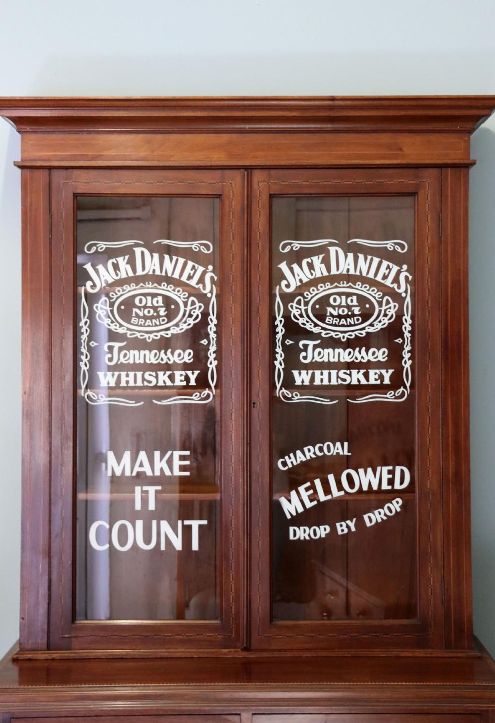A late Victorian warm brown wooden cabinet featuring a glazed upper display section with white Jack Daniels typography and two lower panelled doors. View VII