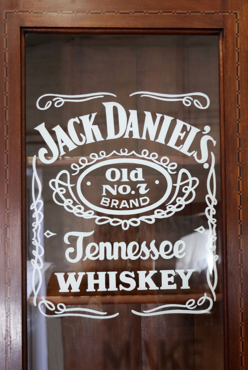 A late Victorian warm brown wooden cabinet featuring a glazed upper display section with white Jack Daniels typography and two lower panelled doors. View XI