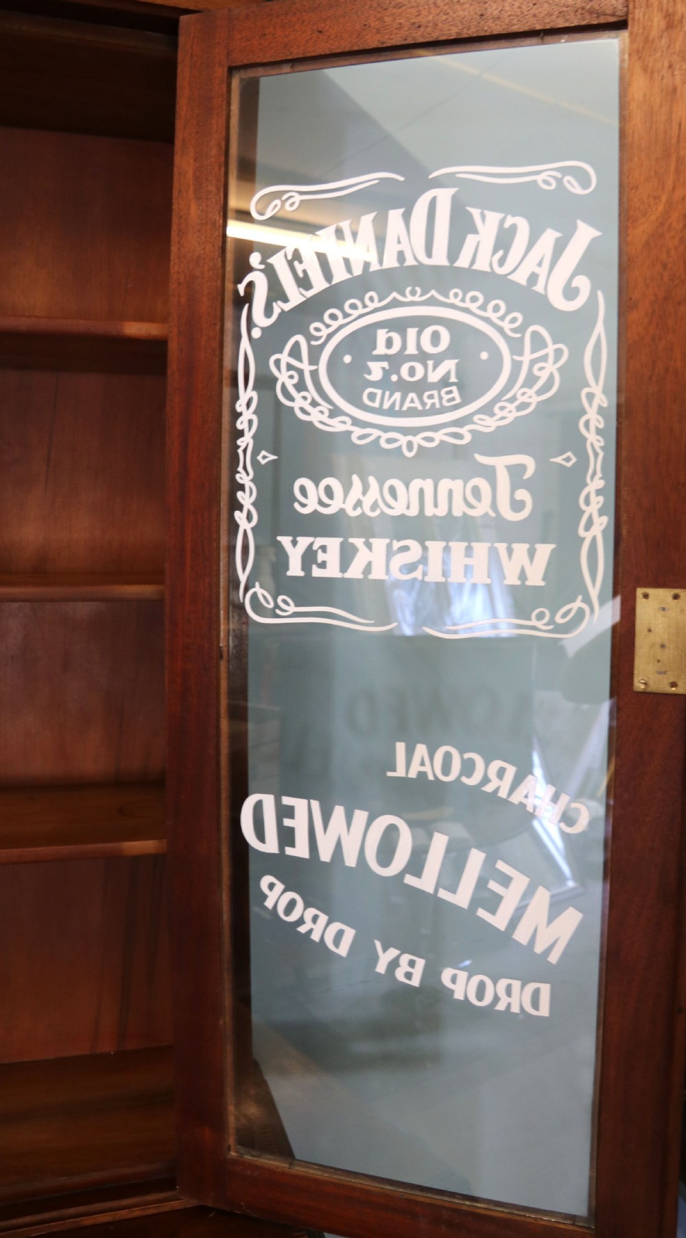 A late Victorian warm brown wooden cabinet featuring a glazed upper display section with white Jack Daniels typography and two lower panelled doors. View XIV