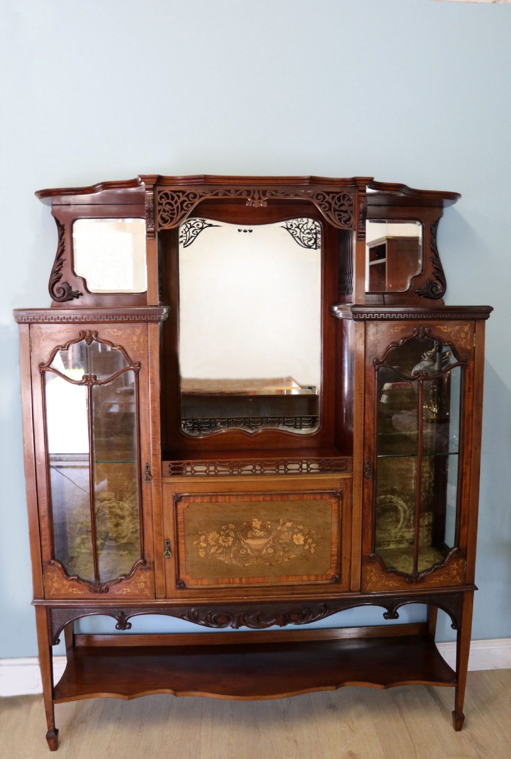 A late 19th-century mahogany display cabinet featuring a drop-centre mirror back, detailed floral marquetry inlays, glazed side doors, and a lower open shelf.