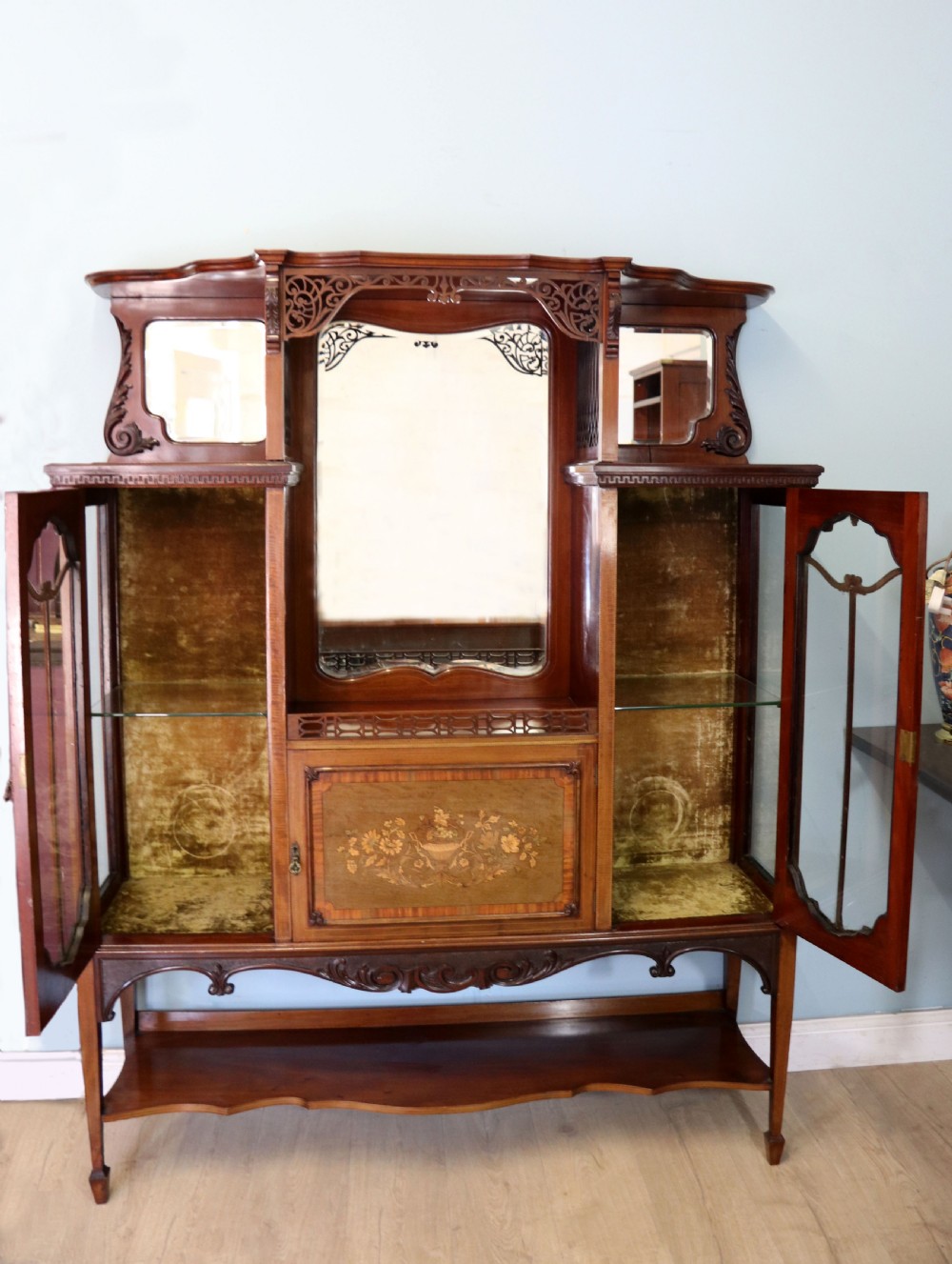 A late 19th-century mahogany display cabinet featuring a drop-centre mirror back, detailed floral marquetry inlays, glazed side doors, and a lower open shelf. View II