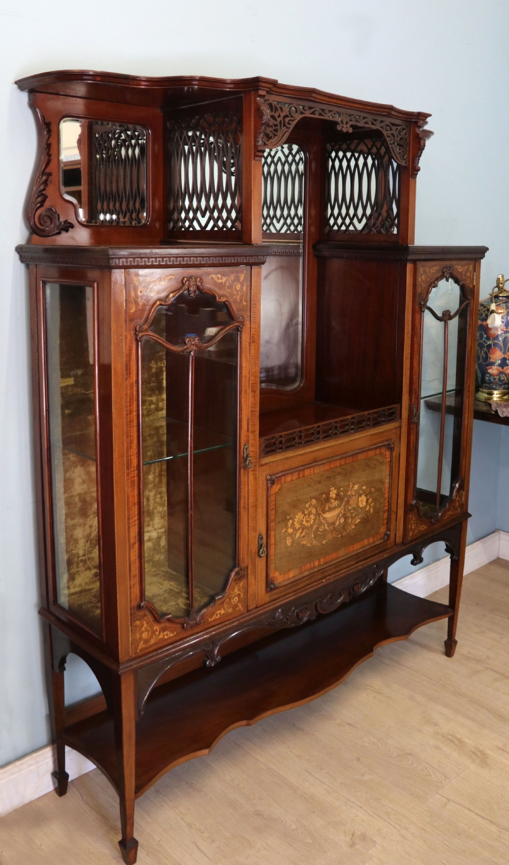 A late 19th-century mahogany display cabinet featuring a drop-centre mirror back, detailed floral marquetry inlays, glazed side doors, and a lower open shelf. View III
