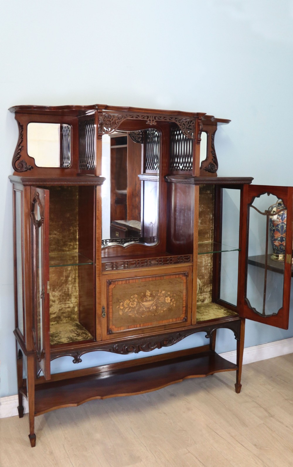 A late 19th-century mahogany display cabinet featuring a drop-centre mirror back, detailed floral marquetry inlays, glazed side doors, and a lower open shelf. View IV