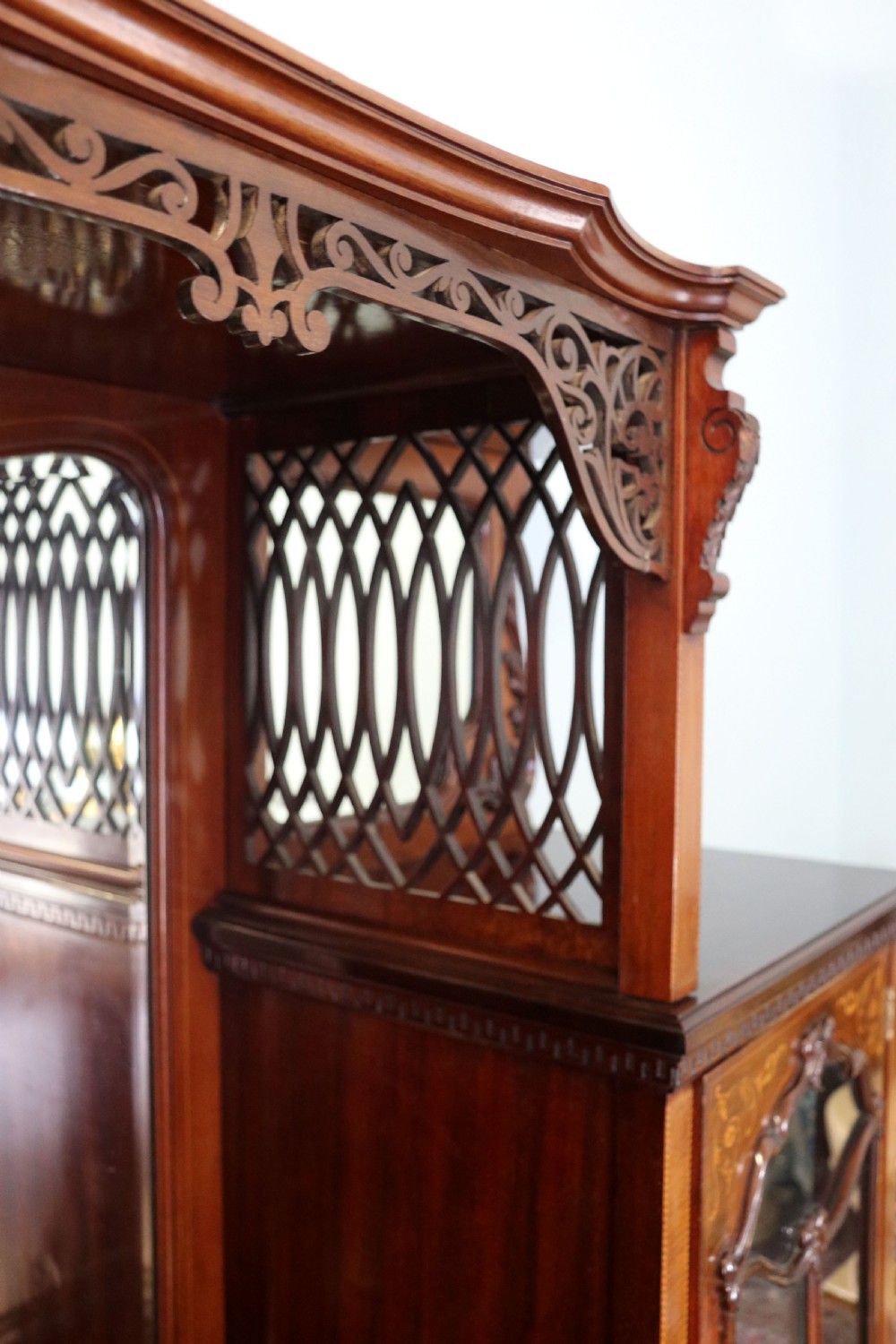 A late 19th-century mahogany display cabinet featuring a drop-centre mirror back, detailed floral marquetry inlays, glazed side doors, and a lower open shelf. View VI