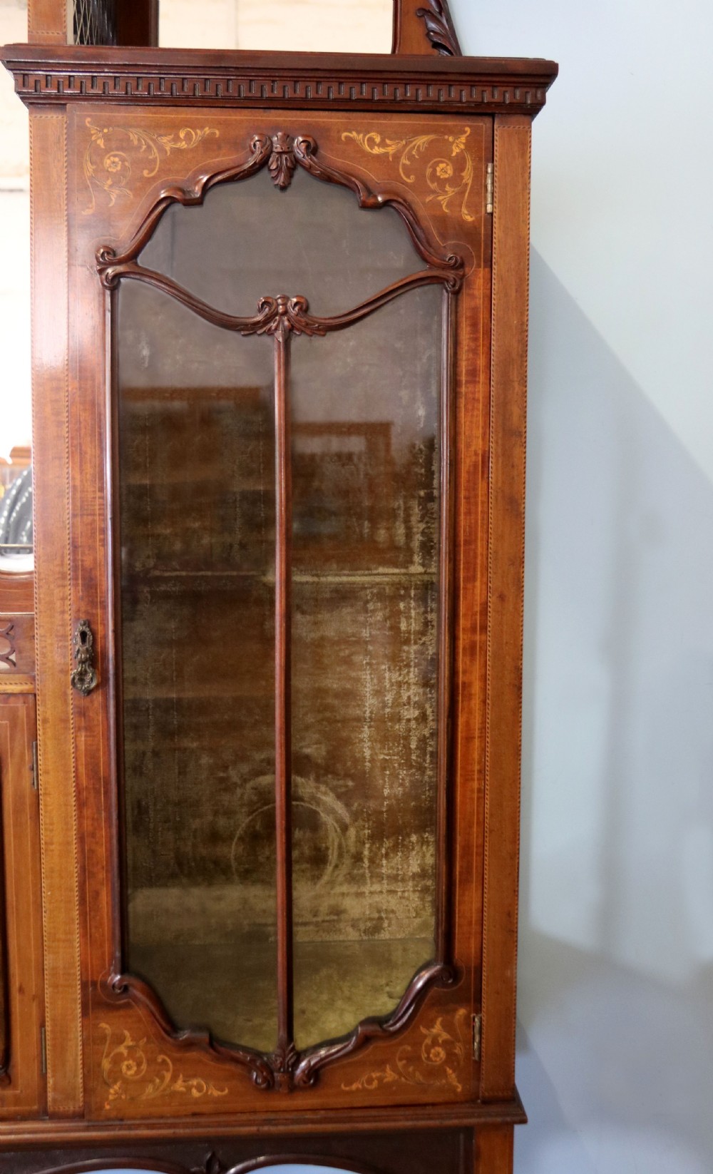 A late 19th-century mahogany display cabinet featuring a drop-centre mirror back, detailed floral marquetry inlays, glazed side doors, and a lower open shelf. View X