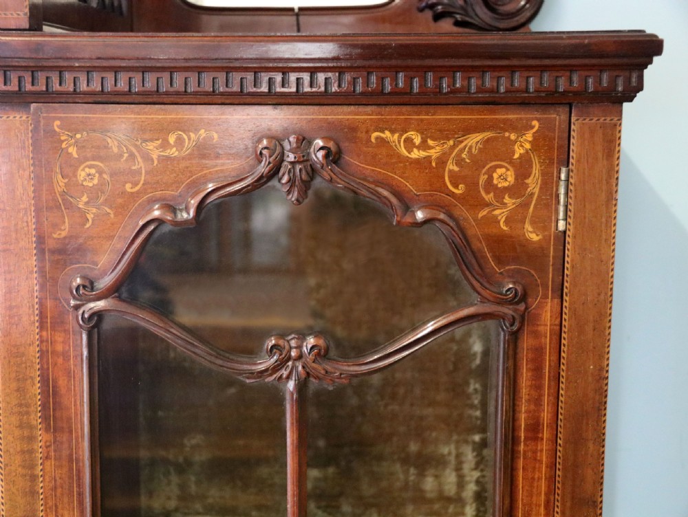 A late 19th-century mahogany display cabinet featuring a drop-centre mirror back, detailed floral marquetry inlays, glazed side doors, and a lower open shelf. View XII