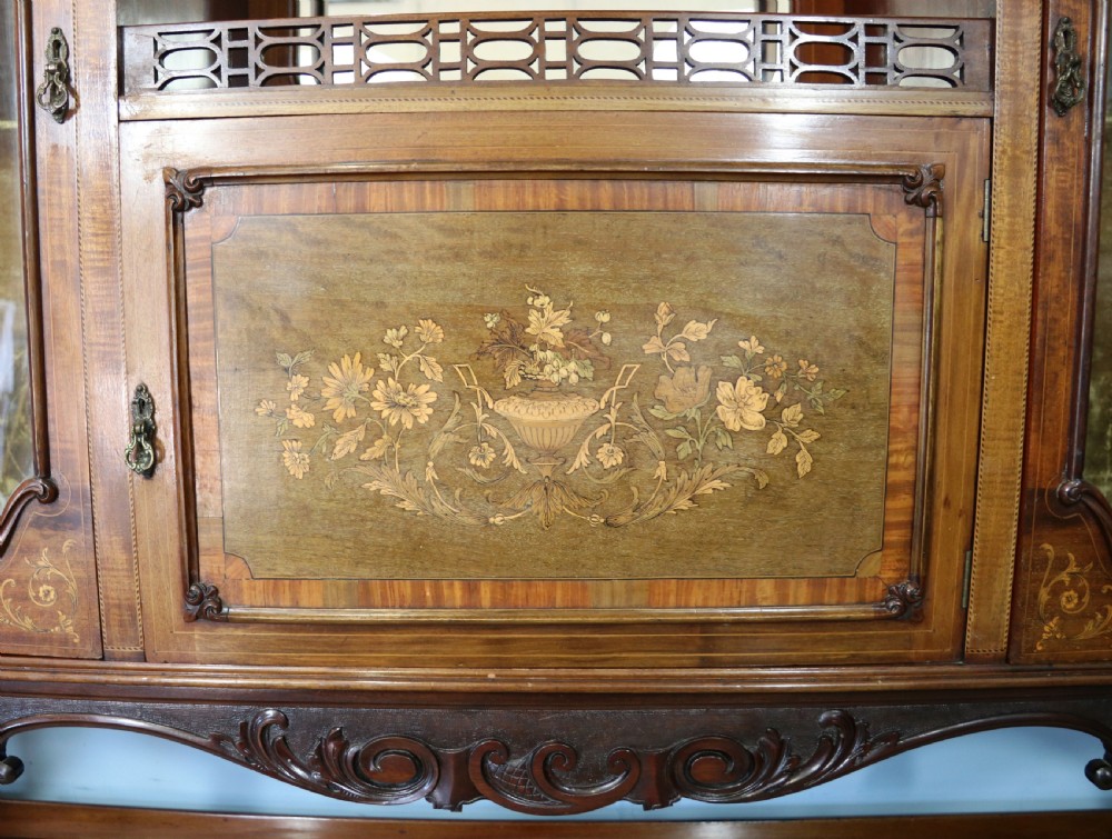 A late 19th-century mahogany display cabinet featuring a drop-centre mirror back, detailed floral marquetry inlays, glazed side doors, and a lower open shelf. View XV