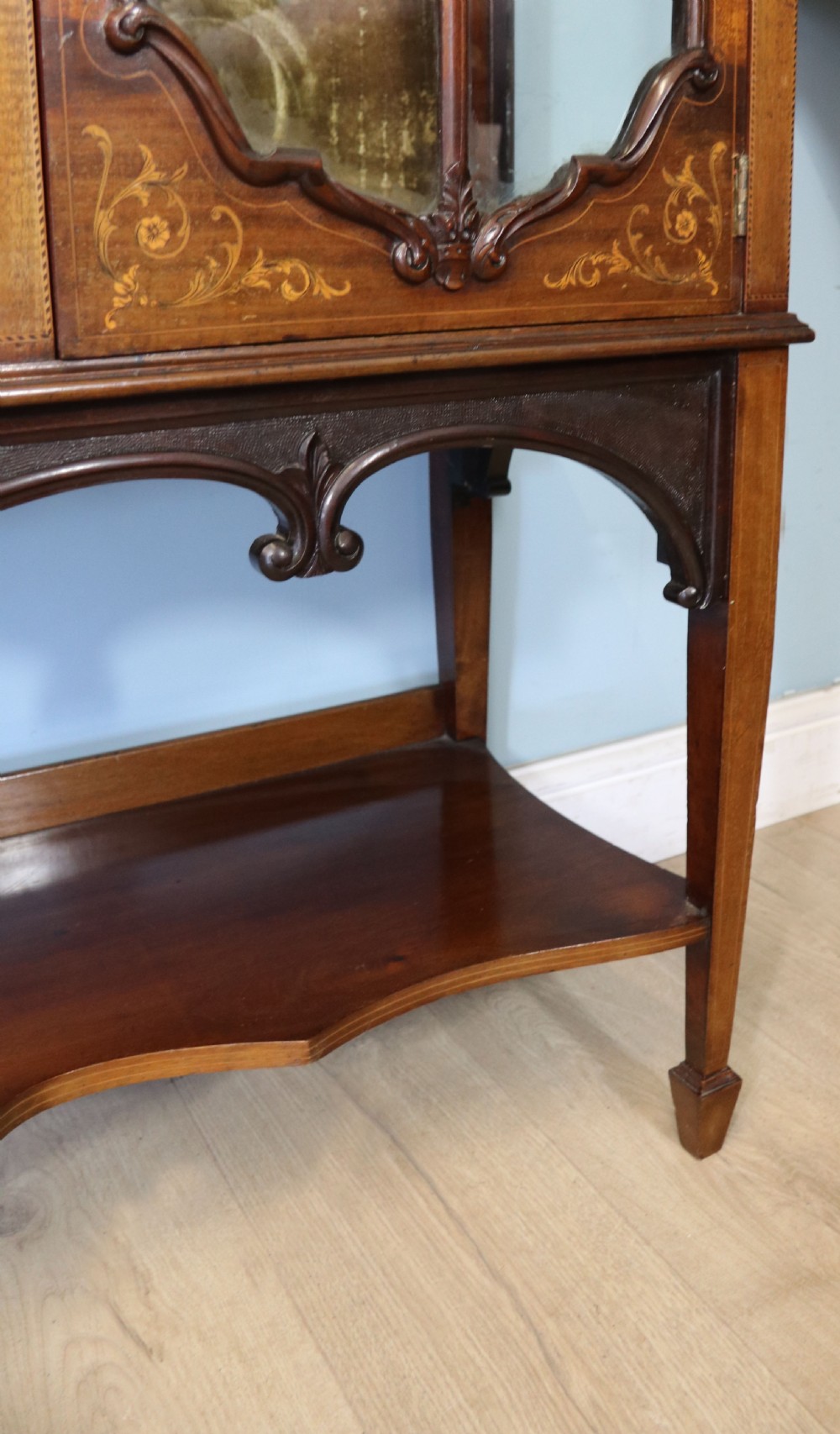 A late 19th-century mahogany display cabinet featuring a drop-centre mirror back, detailed floral marquetry inlays, glazed side doors, and a lower open shelf. View XVIII