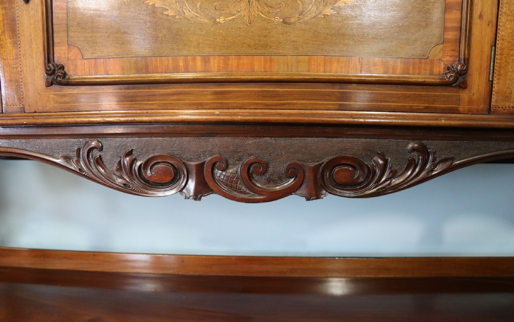 A late 19th-century mahogany display cabinet featuring a drop-centre mirror back, detailed floral marquetry inlays, glazed side doors, and a lower open shelf. View XX