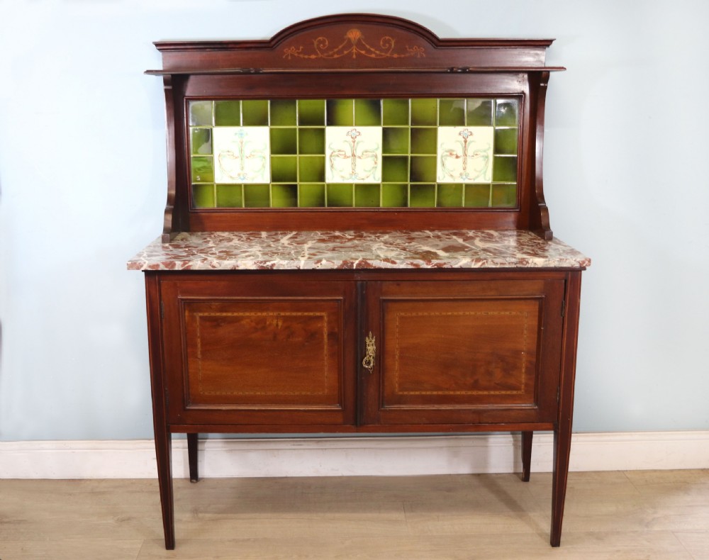 An Edwardian mahogany washstand featuring a red variegated marble top, a splashback set with green ceramic tiles, inlaid cupboard doors, and tapered legs.
