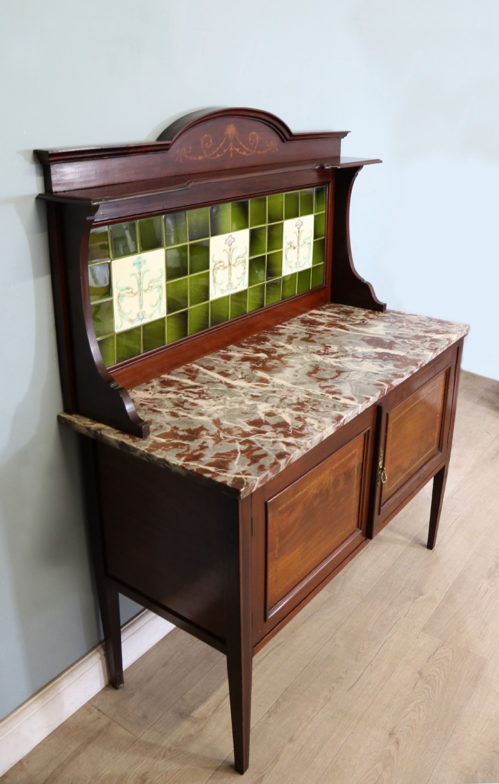 An Edwardian mahogany washstand featuring a red variegated marble top, a splashback set with green ceramic tiles, inlaid cupboard doors, and tapered legs. View V