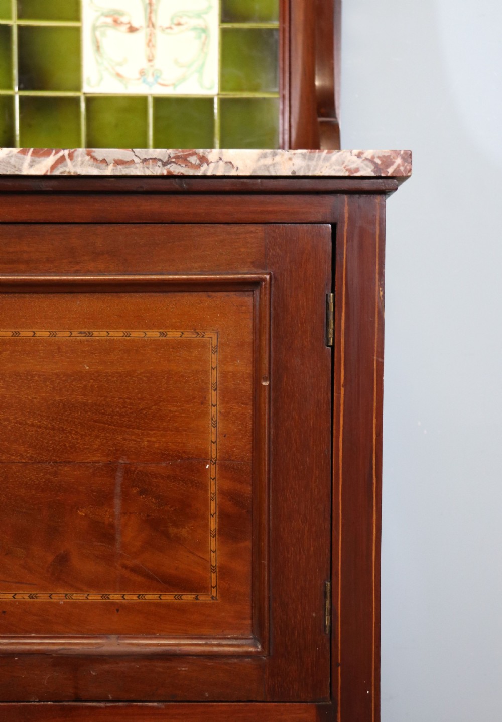 An Edwardian mahogany washstand featuring a red variegated marble top, a splashback set with green ceramic tiles, inlaid cupboard doors, and tapered legs. View XVI
