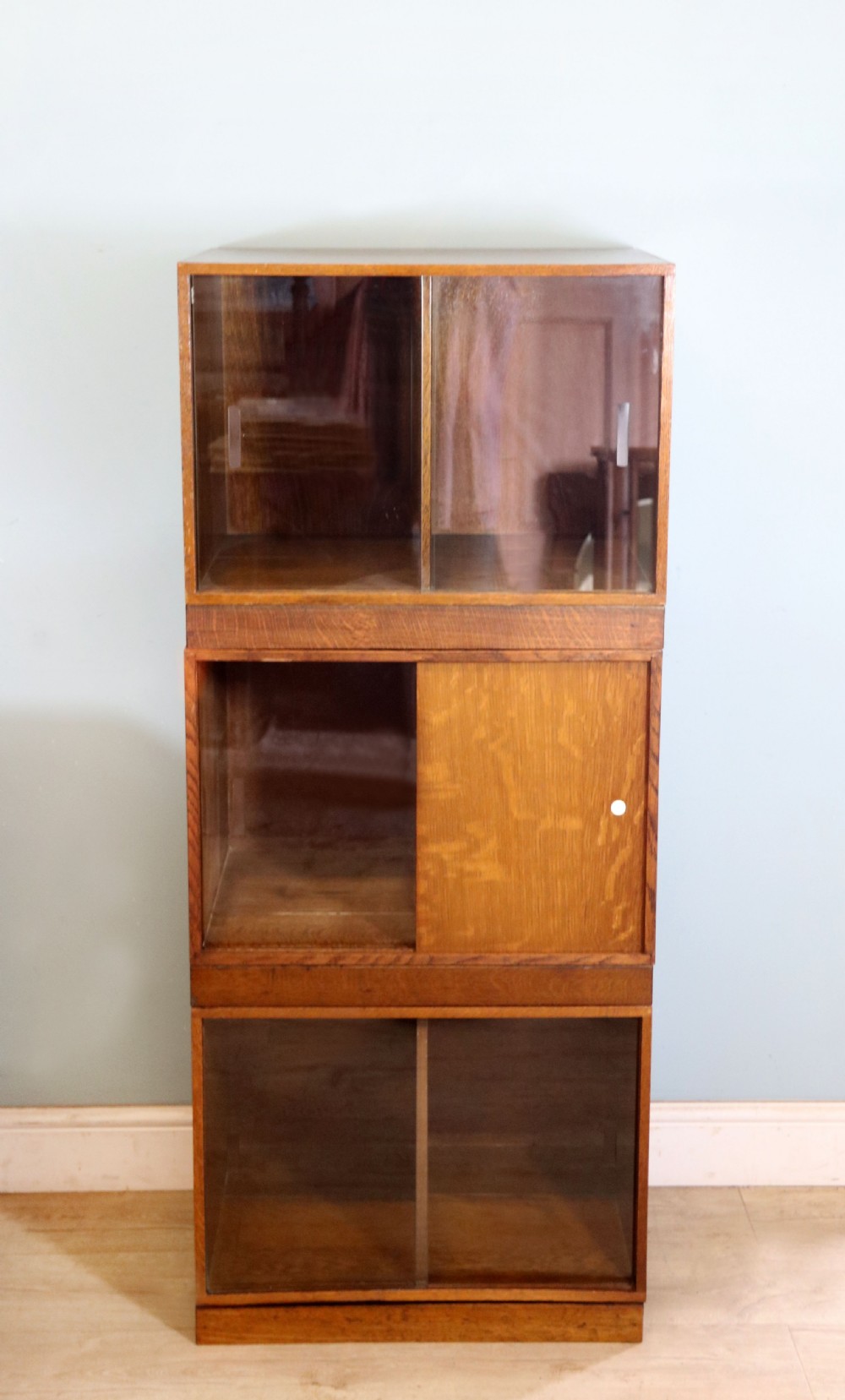 A three-tier mid-century oak sectional bookcase featuring sliding glass doors and a central section with a solid wood panel.