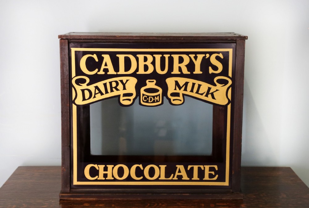 A dark hardwood rectangular shop display cabinet featuring gold lettering on the front glass pane reading Cadburys Dairy Milk Chocolate.