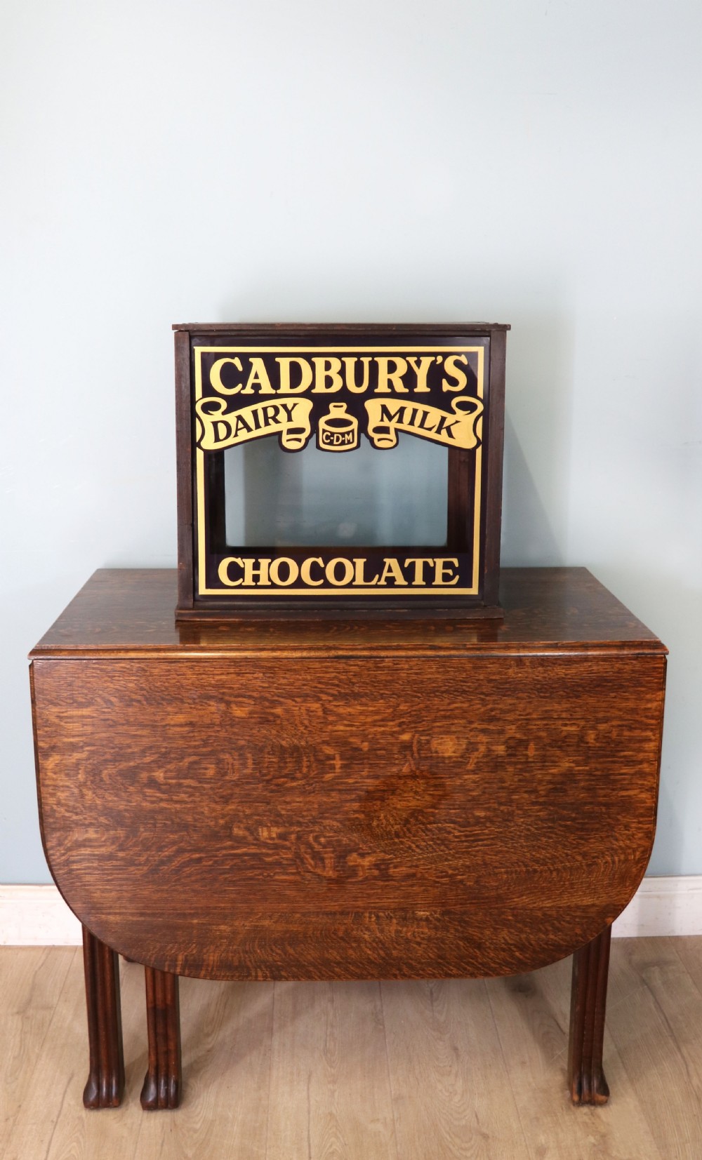 A dark hardwood rectangular shop display cabinet featuring gold lettering on the front glass pane reading Cadburys Dairy Milk Chocolate. View II