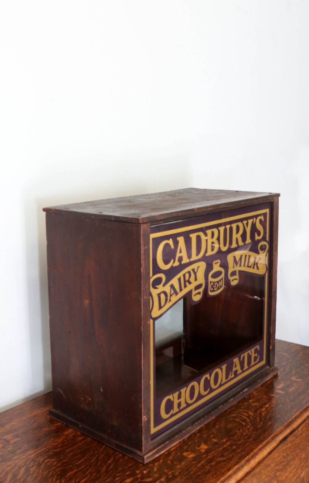 A dark hardwood rectangular shop display cabinet featuring gold lettering on the front glass pane reading Cadburys Dairy Milk Chocolate. View III